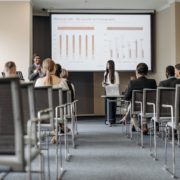 Two women leading a corporate training. They stand holding microphones in front of a projector screen addressing an audience.