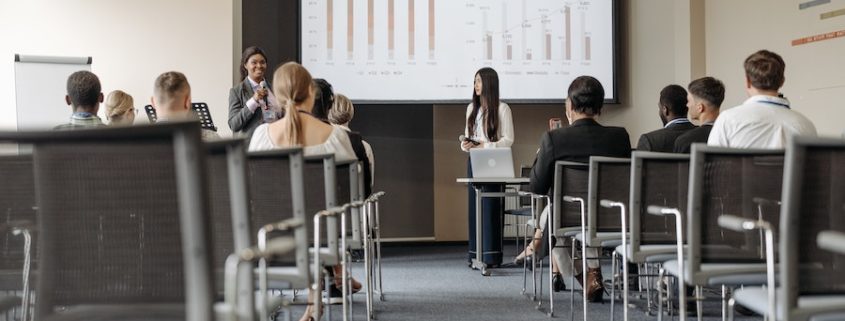 Two women leading a corporate training. They stand holding microphones in front of a projector screen addressing an audience.