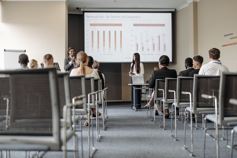 Two women leading a corporate training. They stand holding microphones in front of a projector screen addressing an audience.  