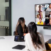 Three people in a conference room look at a display with a Microsoft Teams video meeting.