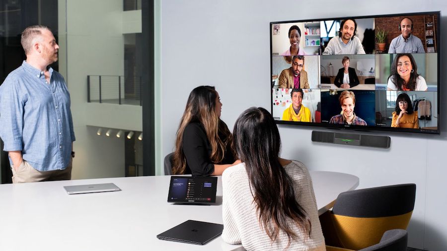 Three people in a conference room look at a display with a Microsoft Teams video meeting.