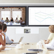 Zoom call being held in an office conference room. Table of attendees is turned to view two screens, one displaying another team and the other screen displaying a graph.