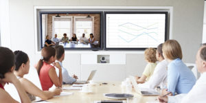 Zoom call being held in an office conference room. Table of attendees is turned to view two screens, one displaying another team and the other screen displaying a graph.