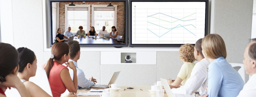 Zoom call being held in an office conference room. Table of attendees is turned to view two screens, one displaying another team and the other screen displaying a graph.