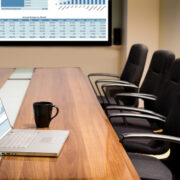 A conference room table that holds an open laptop and a mug with four empty office chairs and a video screen in the background.