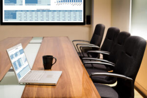 A conference room table that holds an open laptop and a mug with four empty office chairs and a video screen in the background.