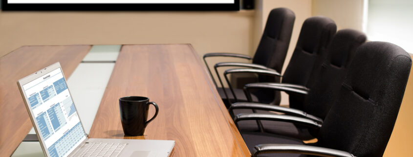 A conference room table that holds an open laptop and a mug with four empty office chairs and a video screen in the background.