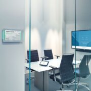 Photo of an empty huddle room, with four chairs placed around a table, a television on the wall, and a smart thermostat outside the room.