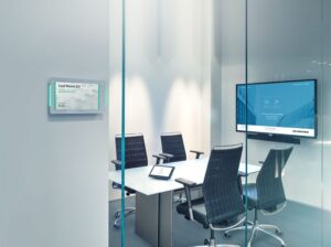 Photo of an empty huddle room, with four chairs placed around a table, a television on the wall, and a smart thermostat outside the room. 