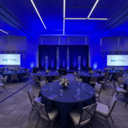 An empty ballroom with several tables and chairs set up for an event with two presentation screens on either side of the stage.