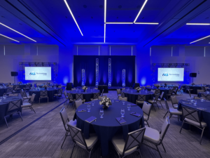 An empty ballroom with several tables and chairs set up for an event with two presentation screens on either side of the stage.