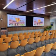 large corporate training room with orange seating in rows facing a podium and double large-screen displays