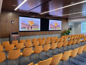 large corporate training room with orange seating in rows facing a podium and double large-screen displays