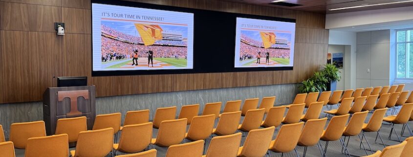 large corporate training room with orange seating in rows facing a podium and double large-screen displays