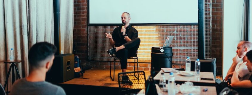 Man on a stage sitting talking into a microphone before an audience.