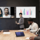 A person is making a presentation in front of a conference room, with remote attendees visible on a display screen.