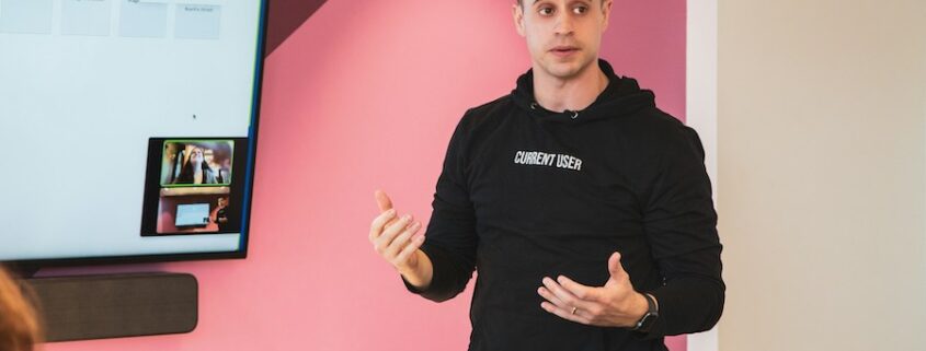 A speaker is explaining a topic in a presentation setting, standing in front of a display screen.