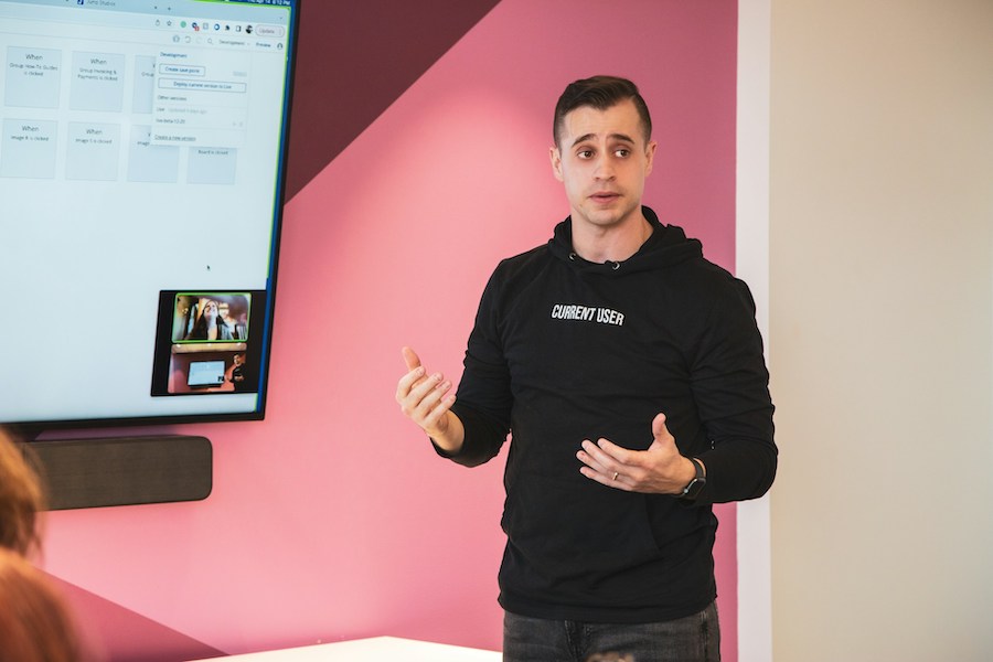 A speaker is explaining a topic in a presentation setting, standing in front of a display screen.