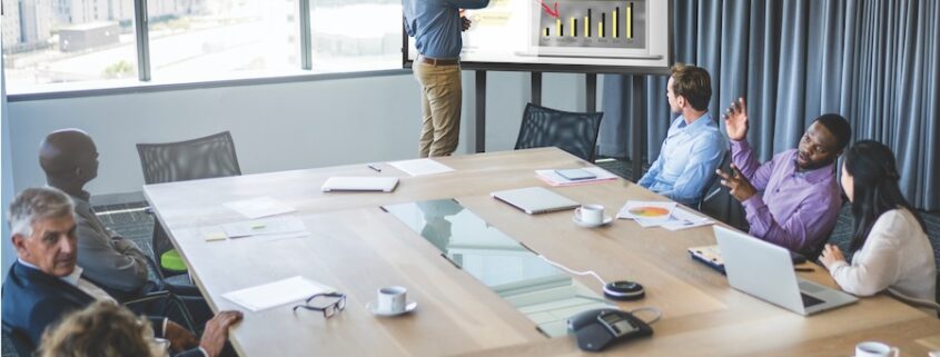 employees sit around a training room table while someone presents on the screen.