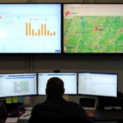 Operator monitoring AV system dashboards and live mapping across five displays in a commercial network operations center.