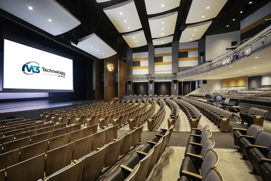 Large auditorium with curved seating, acoustic panels, and an M3 Technology Group display on the stage screen.