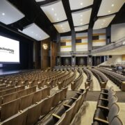 Large auditorium with curved seating, acoustic panels, and an M3 Technology Group display on the stage screen.