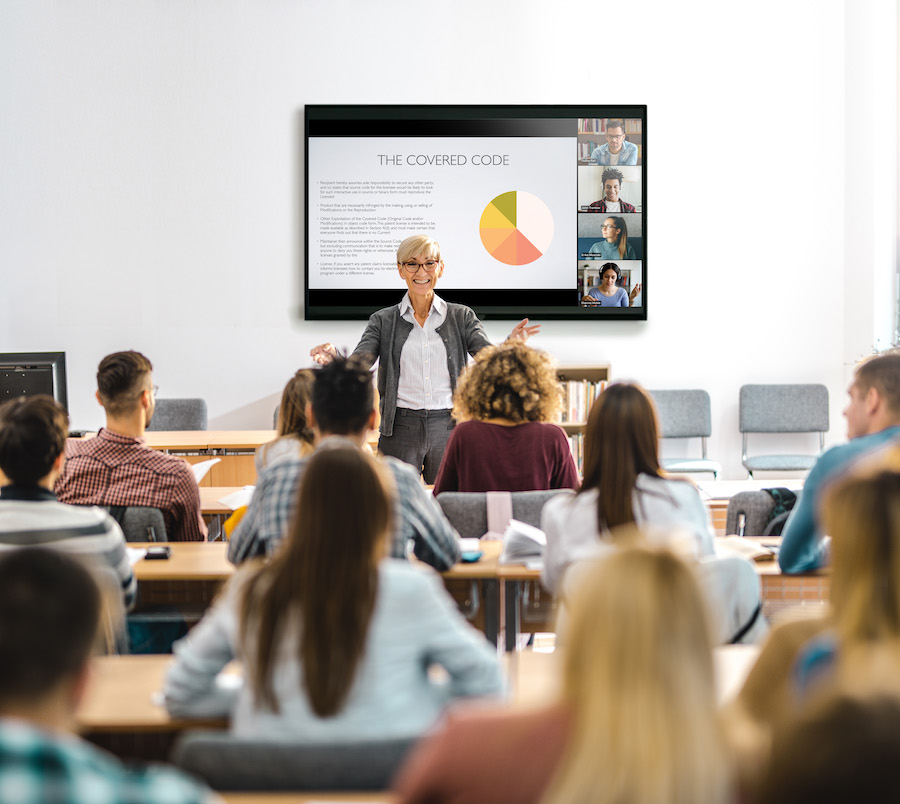 A group of students sitting at long tables with a speaker lecturing in front of them. Behind the speaker, a Crestron screen is mounted on the wall. 