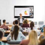 A group of students sitting at long tables with a speaker lecturing in front of them. Behind the speaker, a Crestron screen is mounted on the wall.