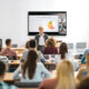 A group of students sitting at long tables with a speaker lecturing in front of them. Behind the speaker, a Crestron screen is mounted on the wall.