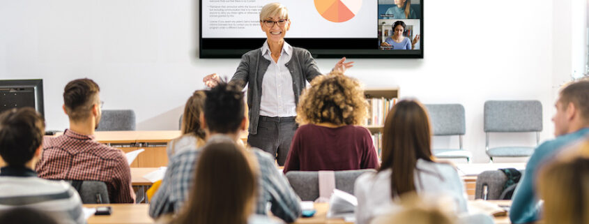 A group of students sitting at long tables with a speaker lecturing in front of them. Behind the speaker, a Crestron screen is mounted on the wall.