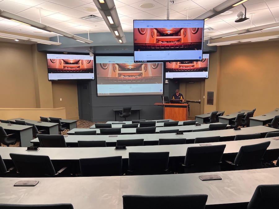 University lecture hall with curved seating and multiple digital displays showing synchronized video content.