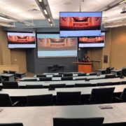 University lecture hall with curved seating and multiple digital displays showing synchronized video content.