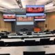 University lecture hall with curved seating and multiple digital displays showing synchronized video content.