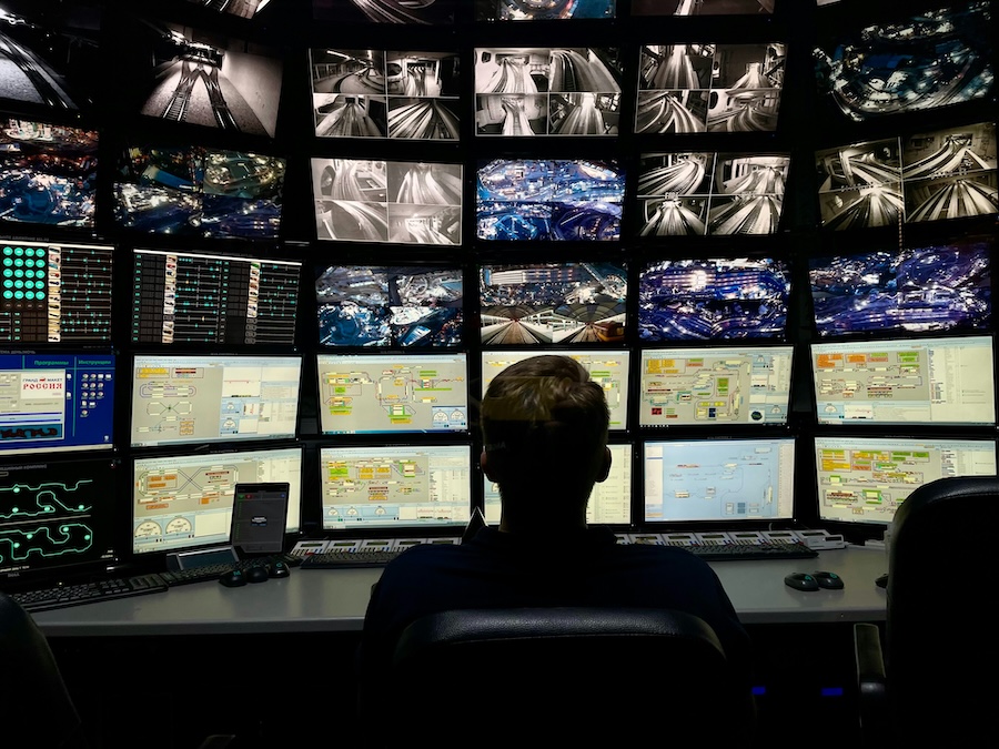 An operator silhouette monitors a large grid of security footage and data dashboards in a dark control room.