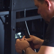 Technician using wire strippers to prep cable near an open AV equipment rack with exposed wiring and hardware.