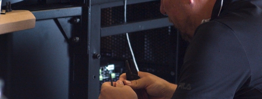 Technician using wire strippers to prep cable near an open AV equipment rack with exposed wiring and hardware.