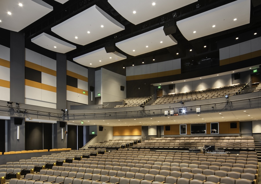 Large tiered teaching theater auditorium with acoustic ceiling panels, stage lighting rig, and rows of empty seating.