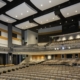 Large tiered teaching theater auditorium with acoustic ceiling panels, stage lighting rig, and rows of empty seating.