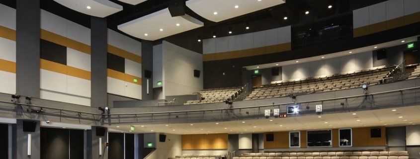 Large tiered teaching theater auditorium with acoustic ceiling panels, stage lighting rig, and rows of empty seating.