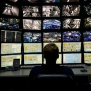 An operator silhouette monitors a large grid of security footage and data dashboards in a dark control room.