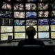 An operator silhouette monitors a large grid of security footage and data dashboards in a dark control room.