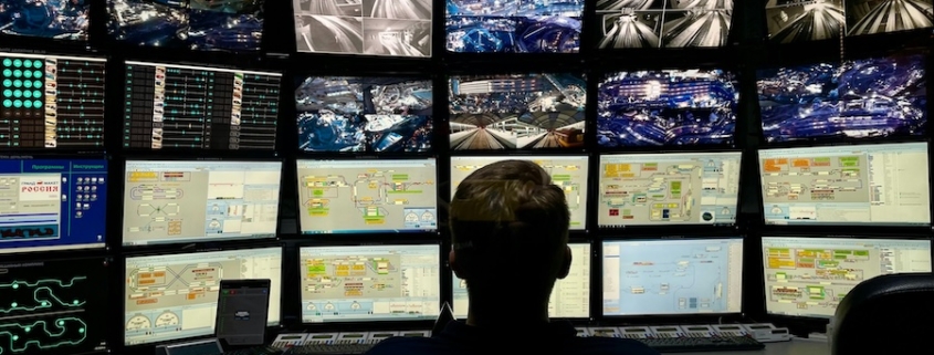 An operator silhouette monitors a large grid of security footage and data dashboards in a dark control room.