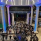 three-story live event space filled with event attendees. Lights are blue-hued and large columns frame the space.