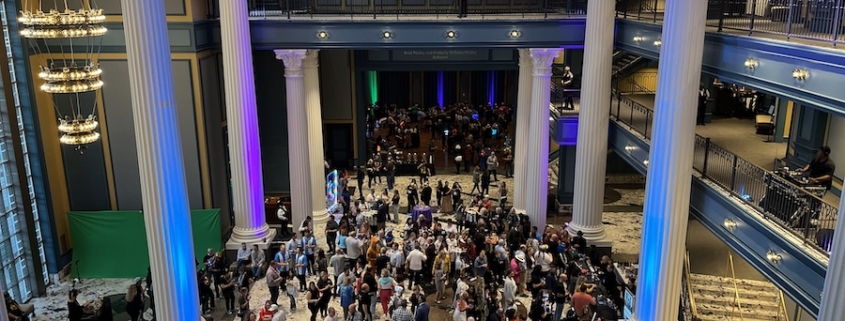 three-story live event space filled with event attendees. Lights are blue-hued and large columns frame the space.
