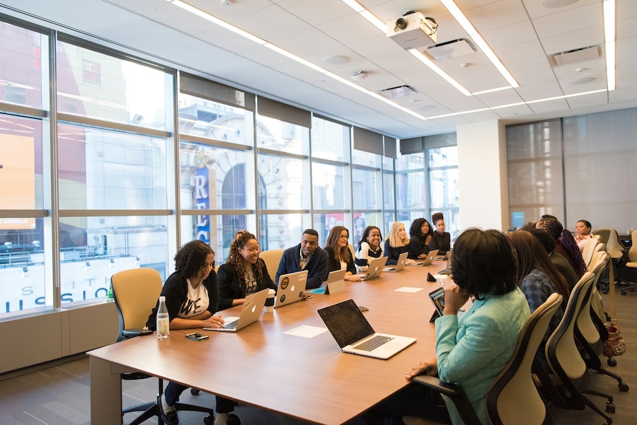 A large group of employees sits around a conference table discussing a meeting agenda item while using laptops