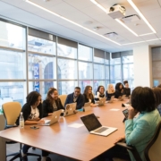 A large group of employees sits around a conference table discussing a meeting agenda item while using laptops.