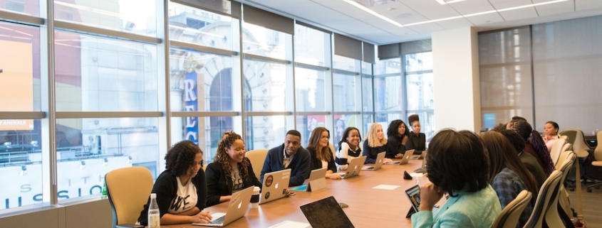 A large group of employees sits around a conference table discussing a meeting agenda item while using laptops.